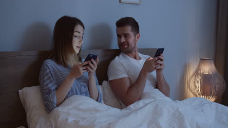 Young-attractive-Caucasian-just-married-man-and-woman-sitting-in-their-bed-late-at-night-and-playing-on-their-smartphones-while-taping,-then-looking-at-each-other.